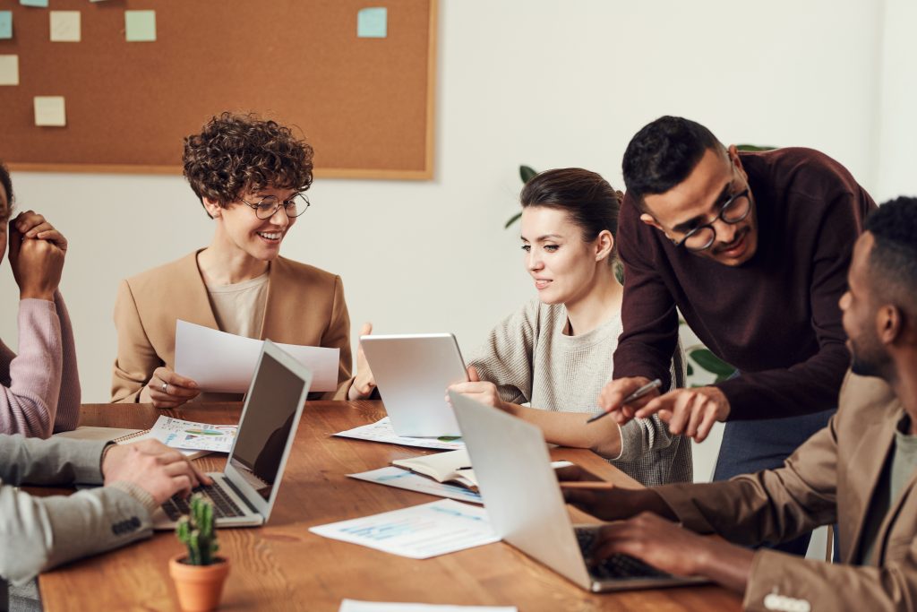 6 employees gather around a table having a meeting on their laptops.