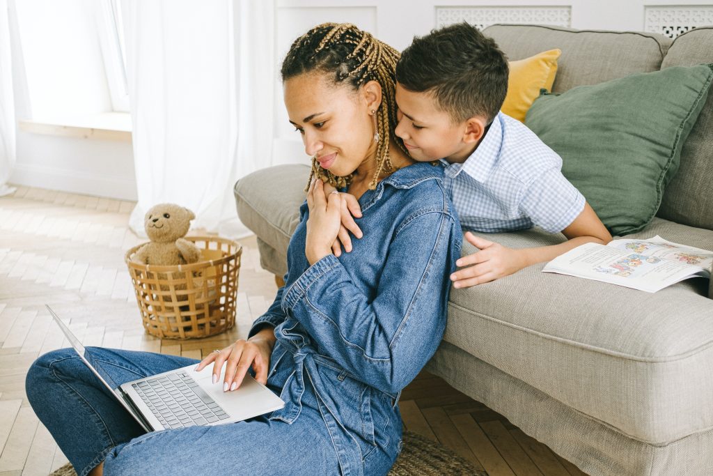 A mother works on her laptop while her son embraces her from behind.