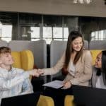 3 smiling employees sit on sofas. 2 of them shake hands.